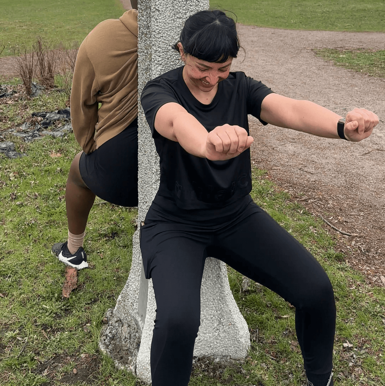 Two members holding a wall sit against a post during a park workout